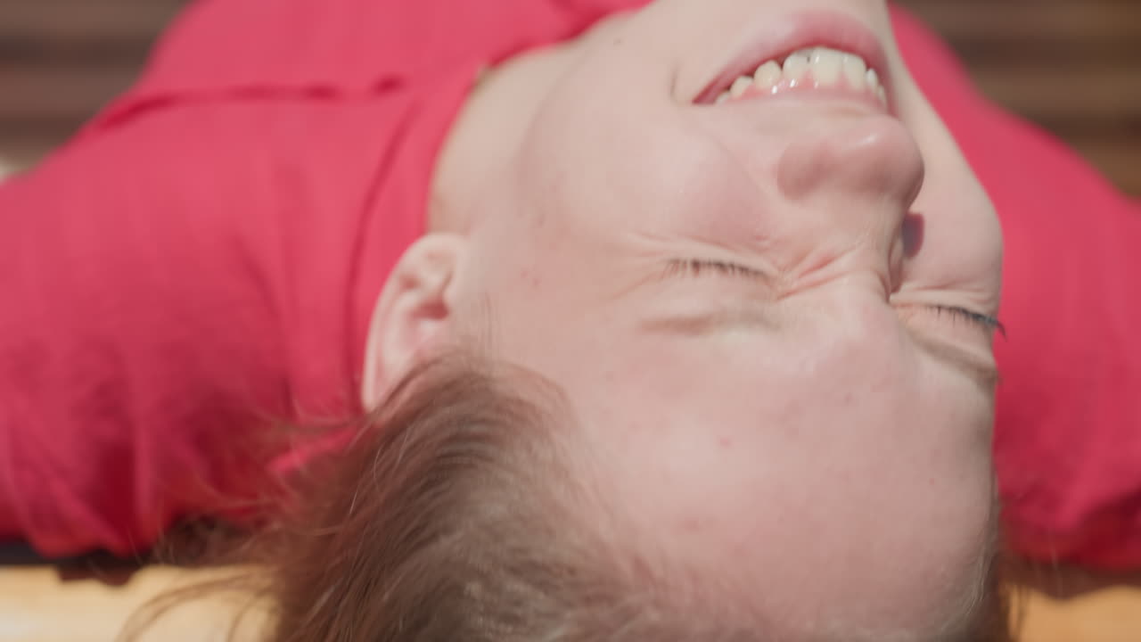 Young woman wearing red top lies on wooden swing bench with eyes closed, sun reflecting off face under bright daylight, surrounded by lush green grass