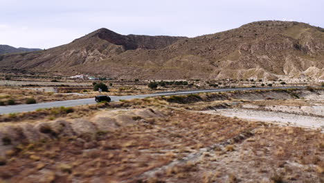 Drone-following-a-black-SUV-driving-over-a-road-in-the-middle-of-a-dry-grassland,-beautiful-mountain-scenery-in-the-background,-Murica,-Spain