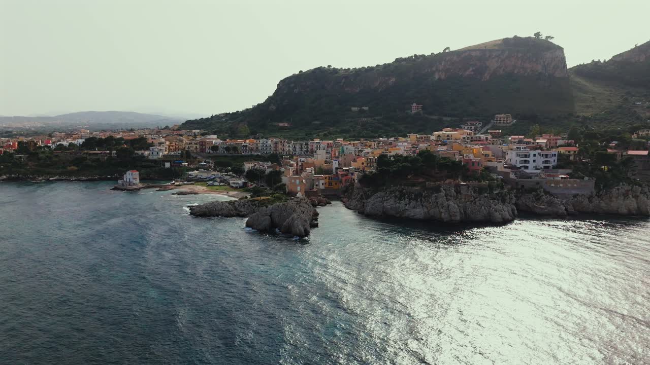 Cinematic drone shot over Porticello in Sicily Italy showing a white lighthouse colorful cliffside homes and Monte Catalfano with the coastline stretching toward Bagheria and Palermo