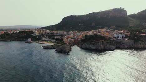 cinematic drone shot over porticello in sicily italy showing a white lighthouse colorful cliffside homes and monte catalfano with the coastline stretching toward bagheria and palermo