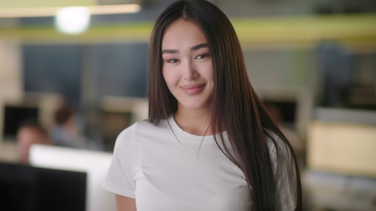 Vista frontal de una hermosa joven profesional con camisa blanca. Retrato de una joven gerente de oficina mirando a cámara y de buen humor. Fondo de oficina borroso