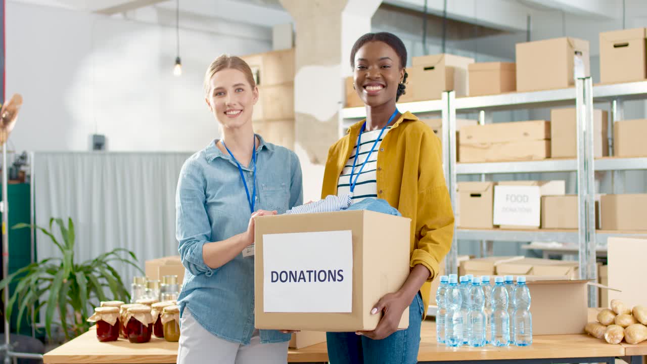 Cheerful young beautiful female volunteers holding donation box with clothes and smiling to the camera in charity warehouse