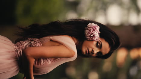 Mujer-Morena-Cautivadora-Vertical-Con-Vestido-Rosa-Con-Flores-En-El-Pelo-Mirando-A-La-Cámara,-Con-Una-Encantadora-Sensación-De-Mareo