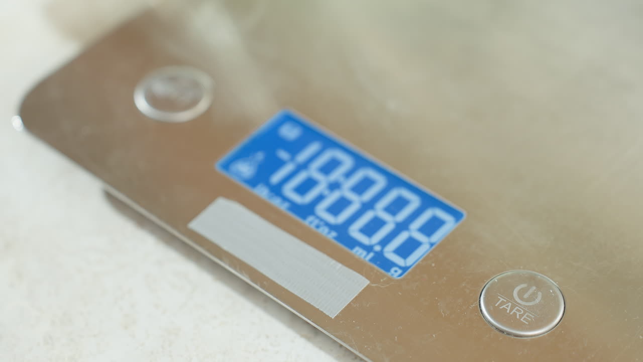 Close up of digital kitchen scale with blue backlit display on countertop as hand reaches over electric stove to test surface temperature, reflecting cautious cooking routine in bright modern kitchen
