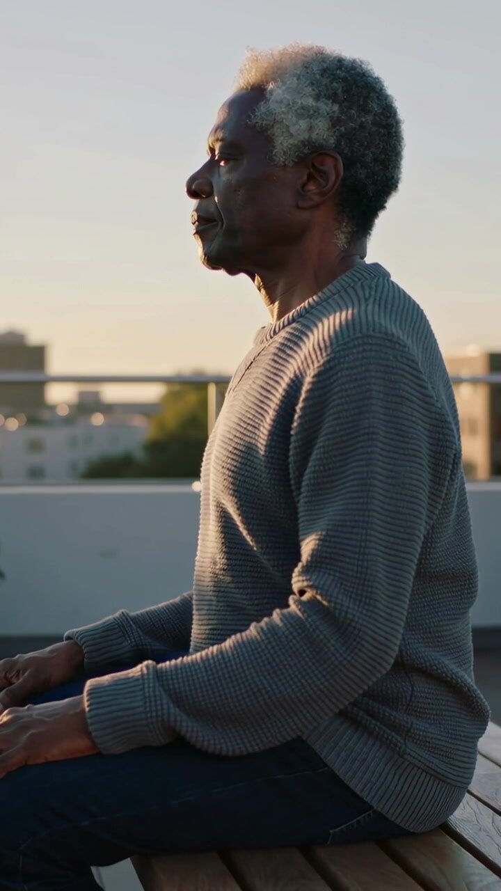 Video verticale: Uomo seduto con un maglione a costine che respira, guarda il tramonto su una panchina sul tetto con una fioriera.
