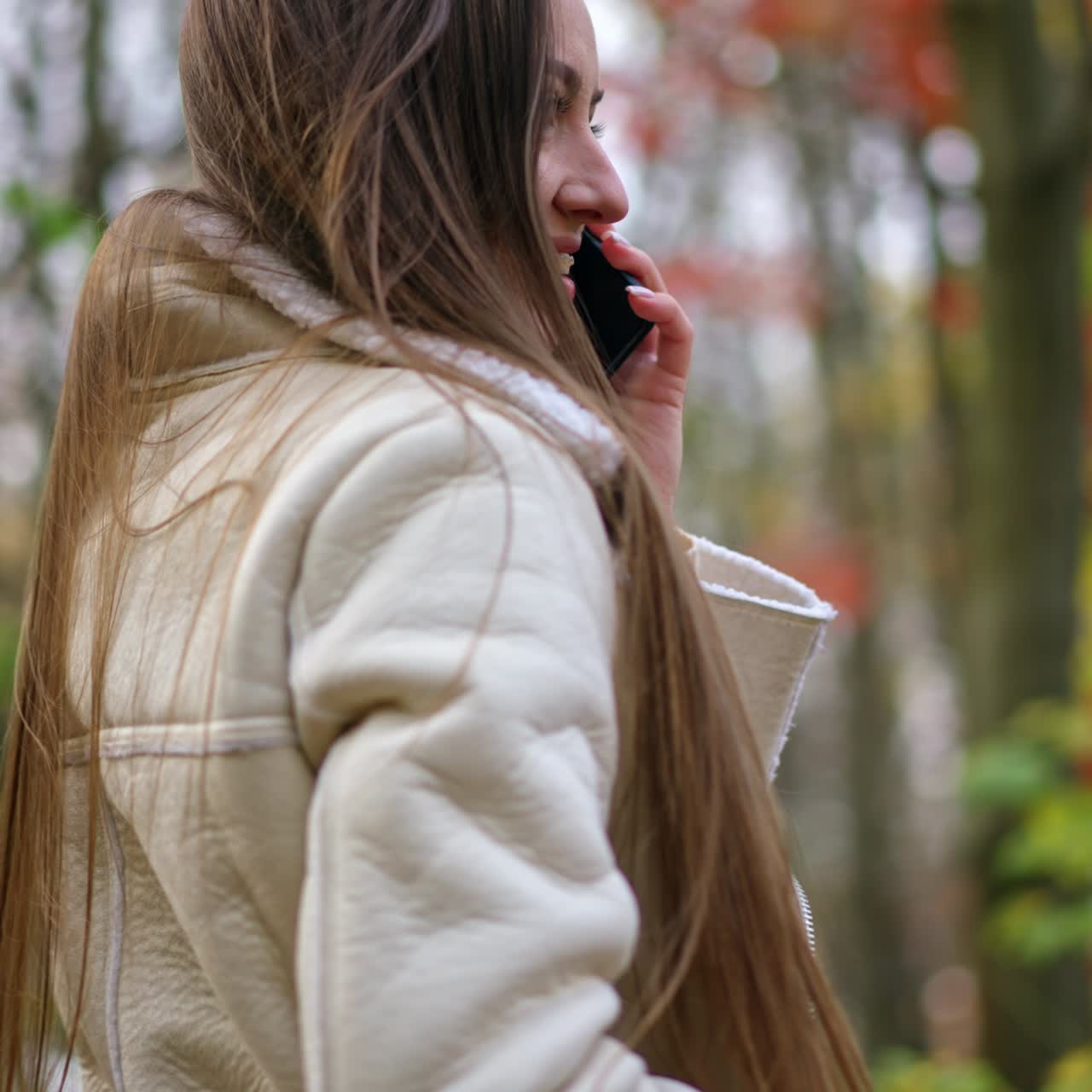 Positive attractive brunette speaking on the phone smiling outdoors. Lady touches her hair looking into camera and holding her phone at the ear