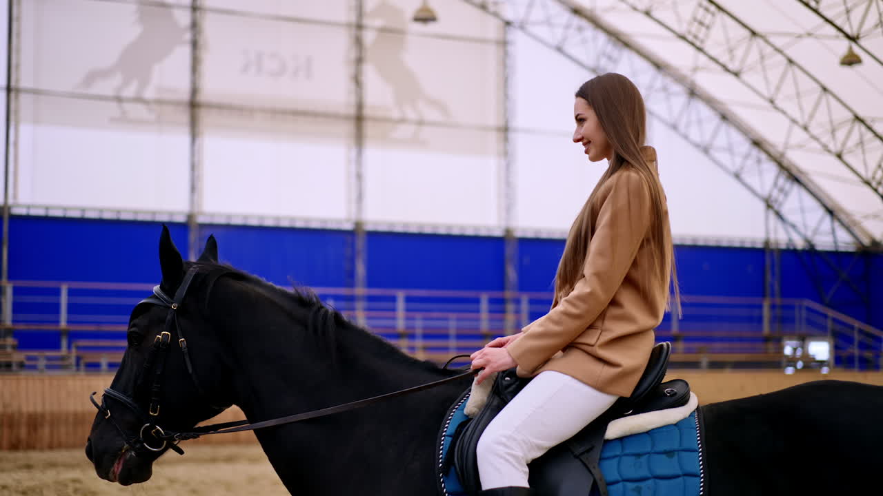 Happy smiling brunette woman riding a beautiful black horse. Lady practicing horse riding in a drill-hall. Blurred backdrop.