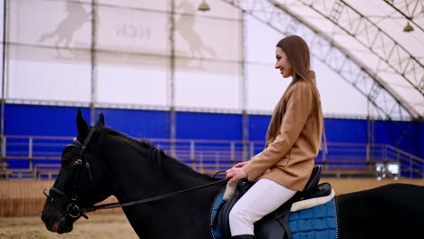 happy smiling brunette woman riding a beautiful black horse. lady practicing horse riding in a drill-hall. blurred backdrop.
