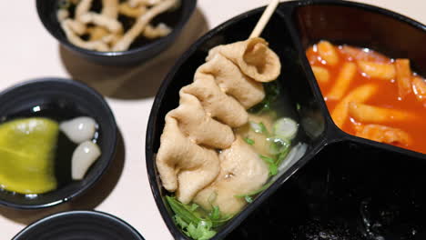 Woman's-hand-pick-up-Odeng-Fish-Cake-on-Wooden-Skewer-from-Plate-with-Broth-and-Eat