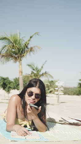 vertical video of happy biracial woman talking on smartphone lying on beach in sun, slow motion