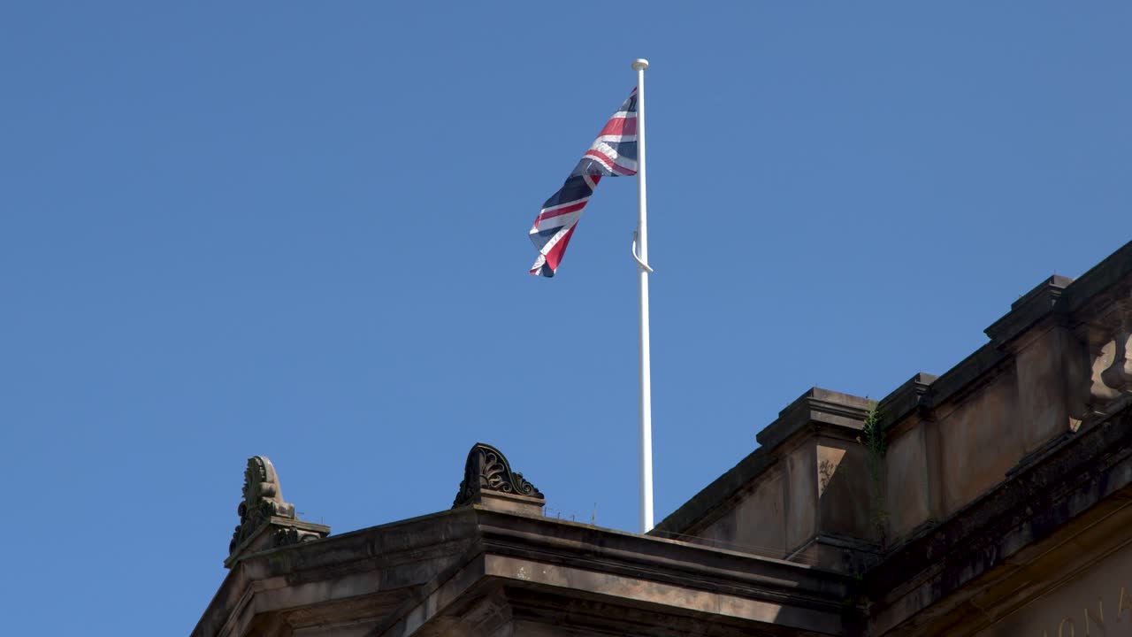 Union flag flutters atop ornate stone building under clear blue sky, midday sunlight, static camera