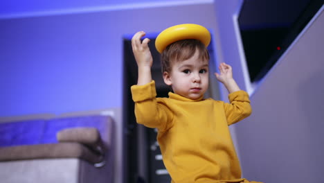 cute baby boy sits still holding a ring from toy pyramid on his head. kid turns his head to shake off the toy. low angle view.