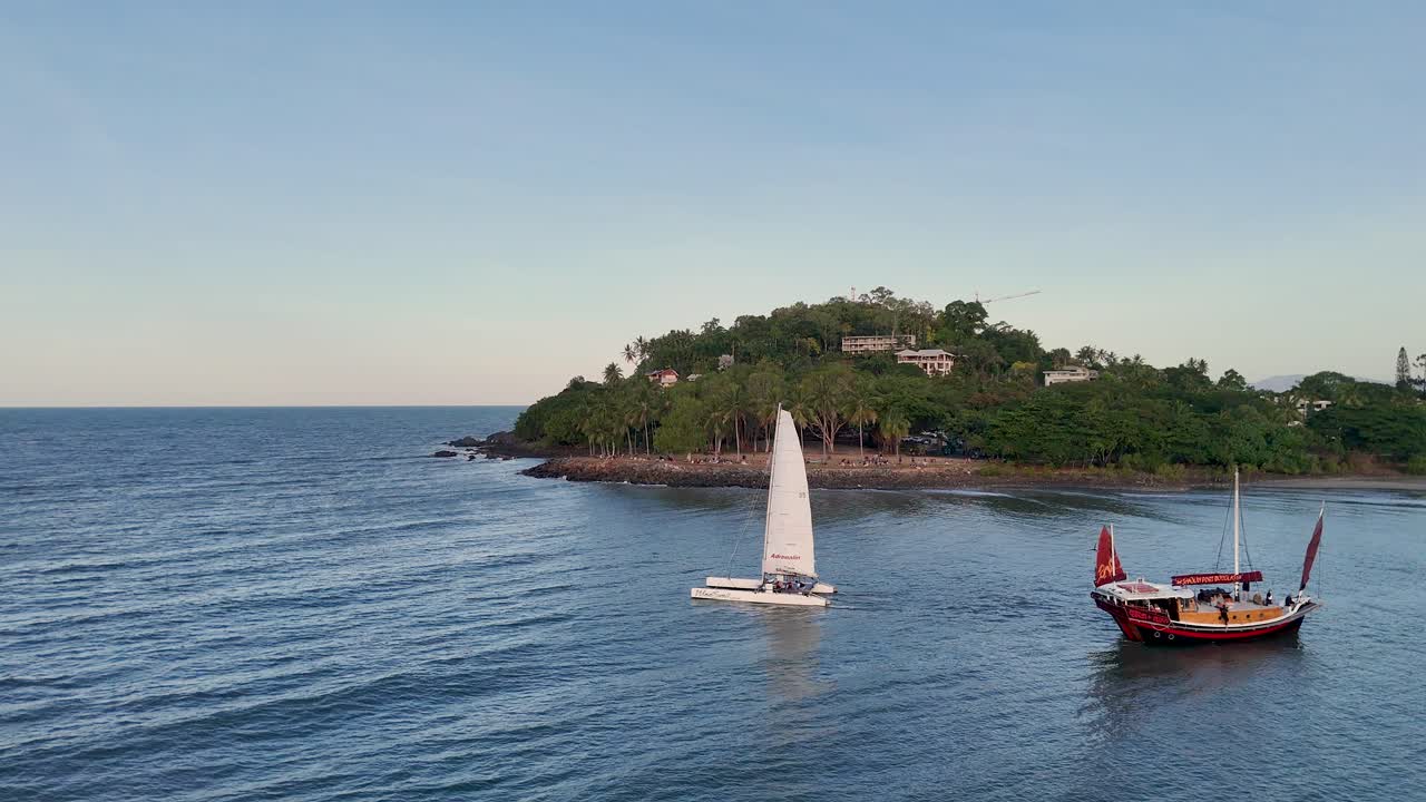 Aerial view of sailboats navigating calm waters near a lush, green island under a clear sky