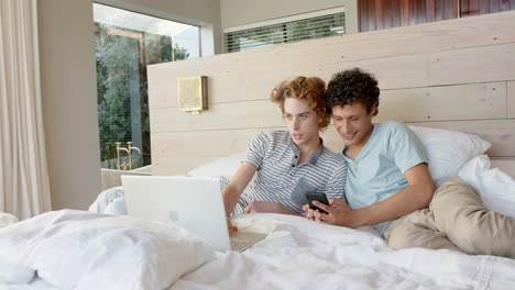 una pareja gay feliz y diversa acostada en la cama y usando un teléfono inteligente y una computadora portátil en casa, en cámara lenta