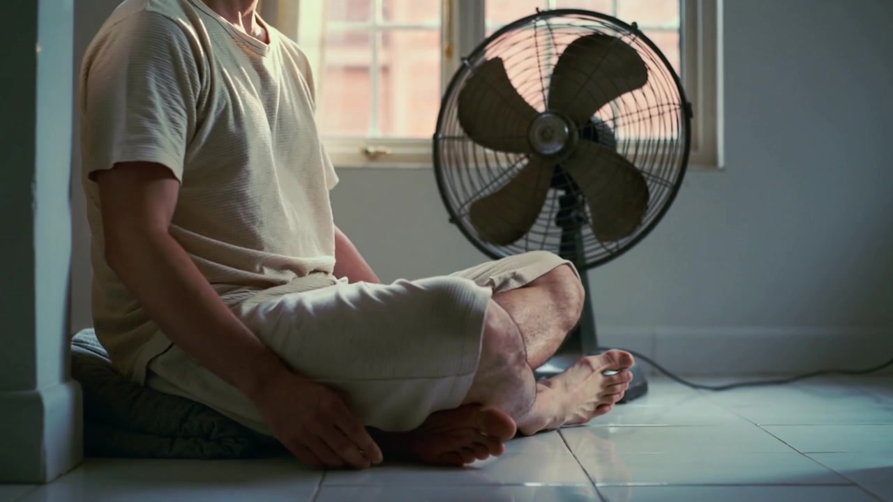 Entering man in light tee and shorts at home, approaching fan, sitting on cushion cooling down