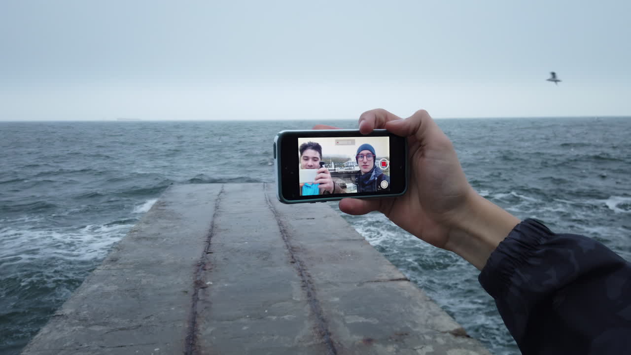 Chisinau, Moldova - November 10, 2021: Two friends smile for a selfie while standing on a pier by the ocean. The cloudy sky and waves create a serene backdrop for their memorable outing