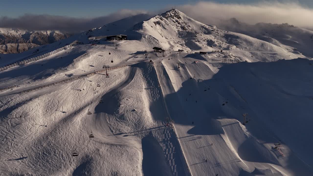 Many Skiing people and tourist on snowy slopes of Cardrona Ski Resort at sunrise. Drone wide shot. Hovering clouds over snowy summit. Ski Lift to base in New Zealand