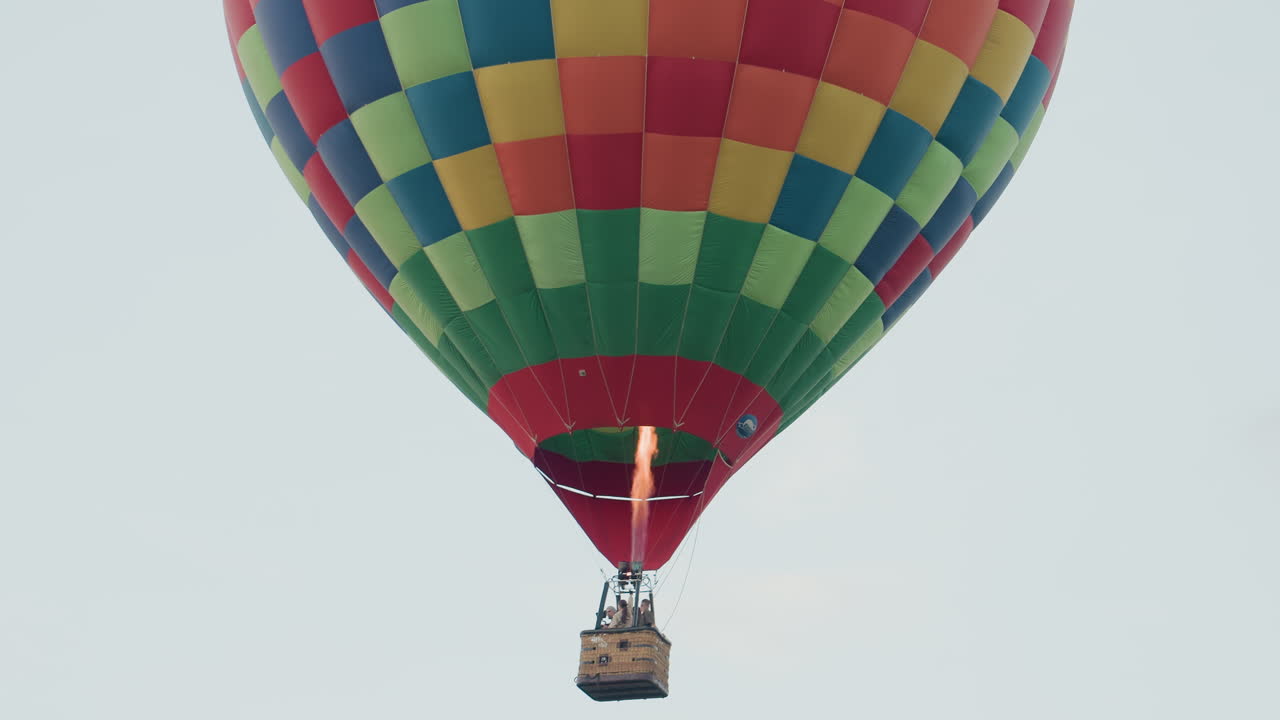 three passengers rising in vibrant checkered hot air balloon high above open green field with distant second balloon drifting on horizon against soft pastel sky