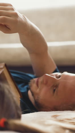 CU, Side view, concept of distance learning: little girl and her bald unshaven dad lie on floor, father knocks on book, which kid covered face, woke up, and shows in notebook on task, closeup