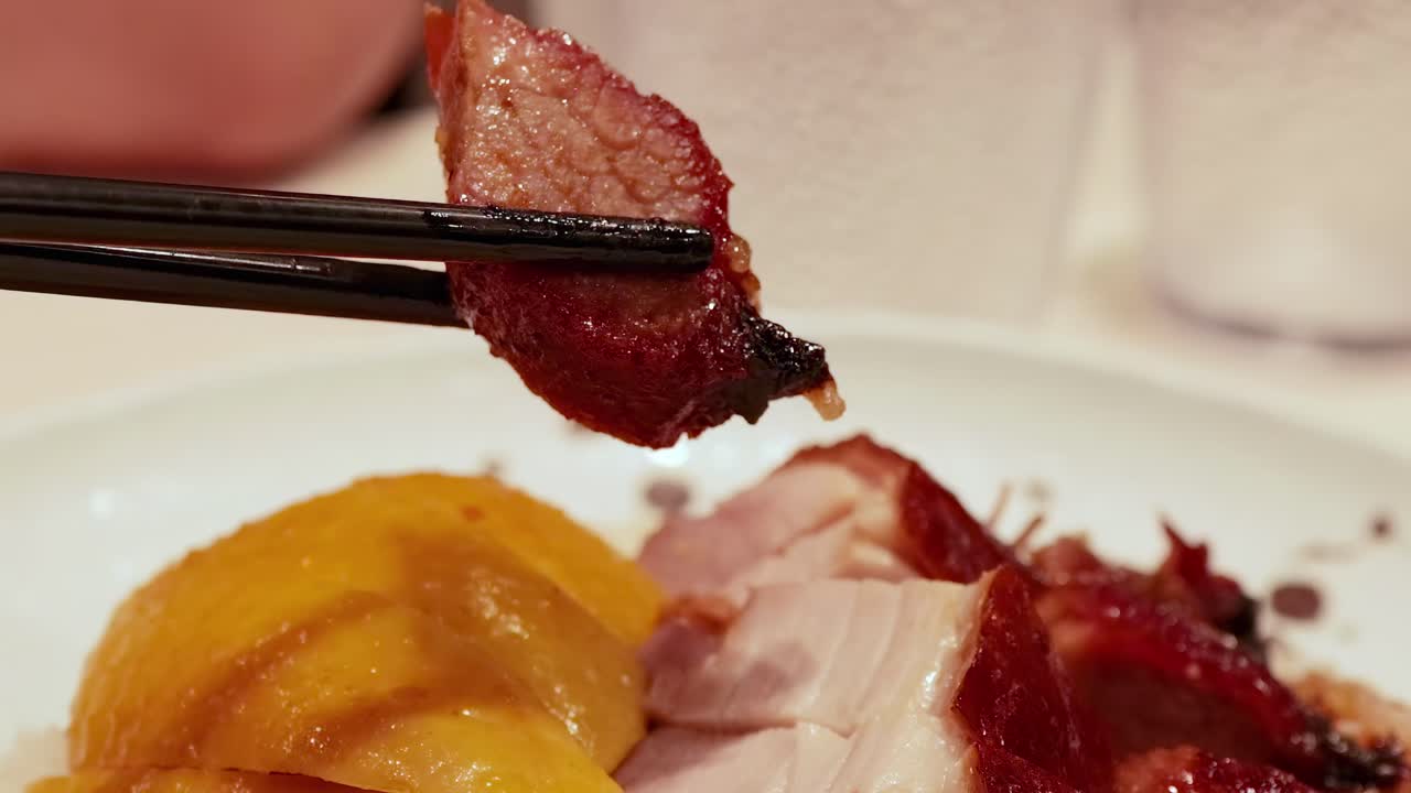 Close-up of chopsticks picking up juicy roast meat pieces from a plate with sliced mango.