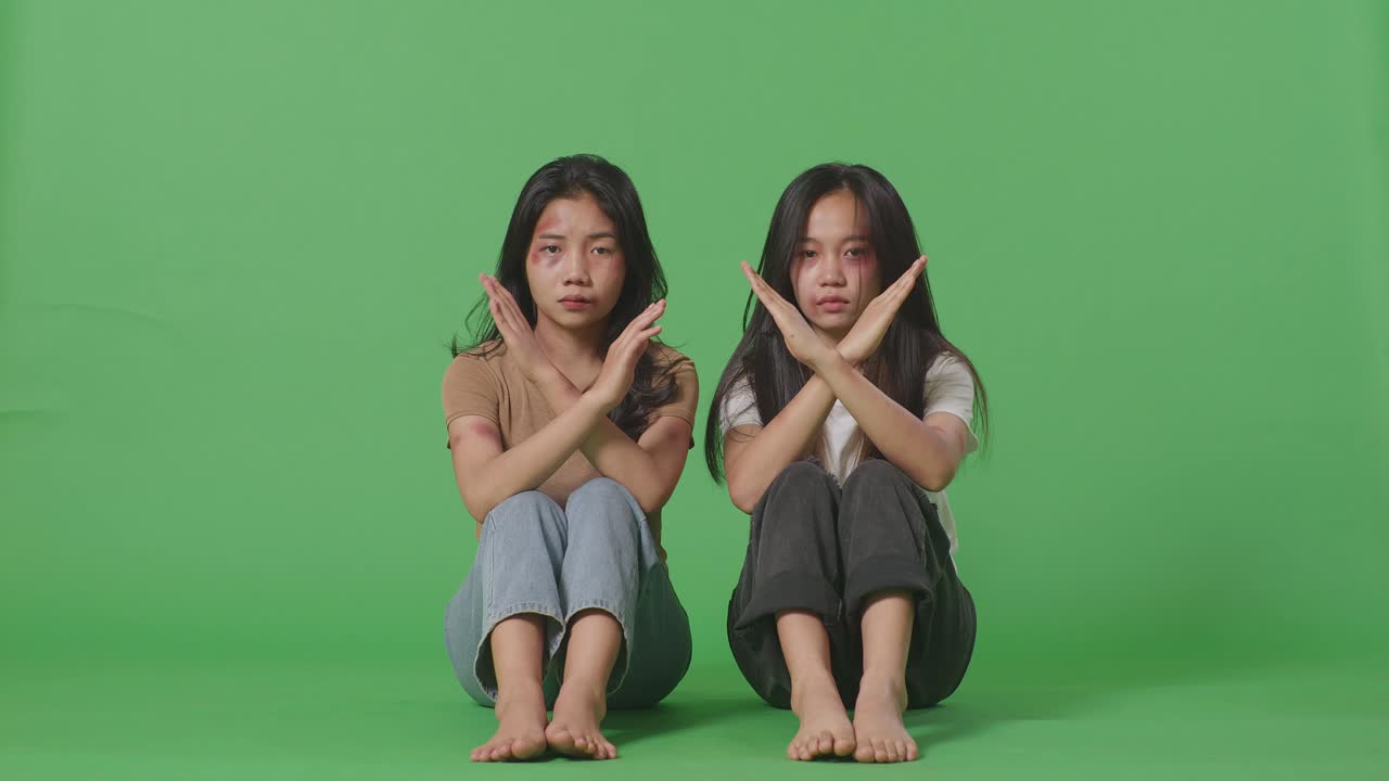 Full Body Of Young Asian Women Victims Of Violence With Bruise On Bodies Looks Into Camera Showing Hands Cross To Stop Violence While Sitting In The Green Screen Background Studio