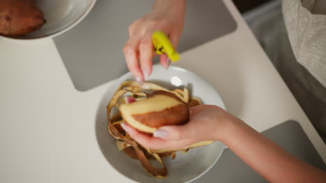 close up overhead view of cook using green-handled peeler to remove skin from sweet potato over white bowl filled with discarded peels on kitchen table with soft lighting and warm tone
