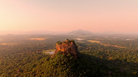 a peaceful sunrise drone shot of sigiriya rock rising above the dense sri lankan jungle, with a calm lake reflecting the first light of day.