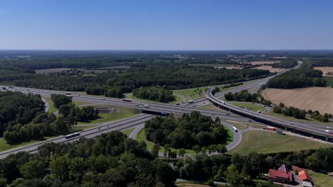 german highway junction in rural area with green forest trees in summer. aerial backwards rising shot. traffic on multi-line autobahn interchange.blue sky and farm houses in countryside.blue road sign