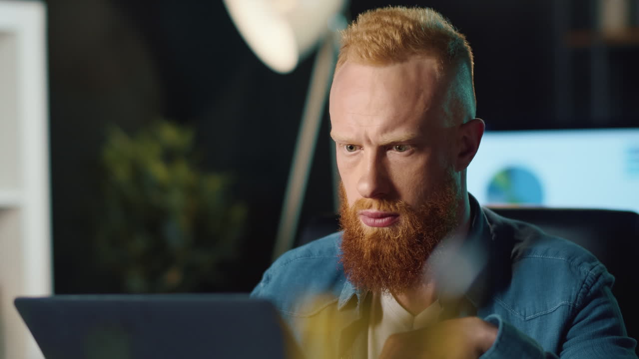 Konzentrierter Hipster-Mann gibt Daten am Computer im Nachtbüro ein. Mann blickt auf den Bildschirm.