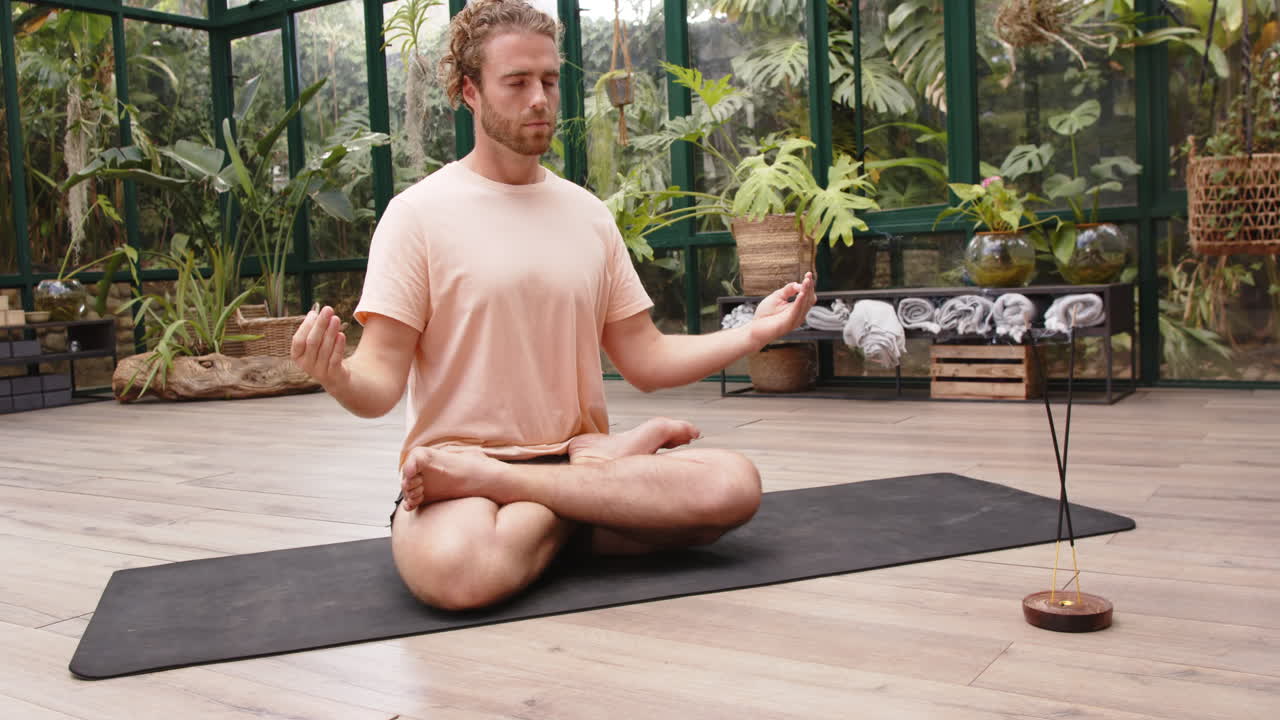 Meditating on yoga mat, man practicing mindfulness in glass house garden