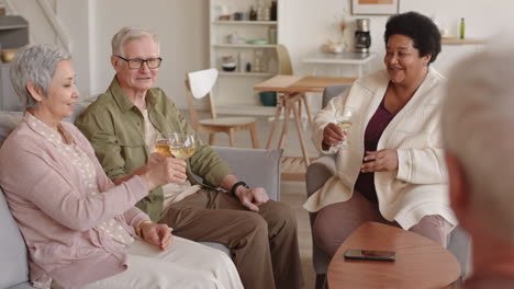 medium long of diverse grey-haired couple sitting on couch, african woman in armchair, talking, looking at unrecognizable friend, drinking white wine together