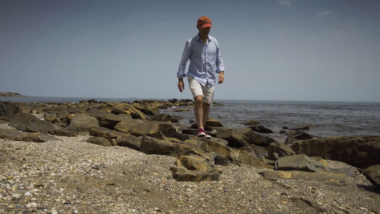 Man Sits on the rocks on the beach front then gets up and walks off past the camera.