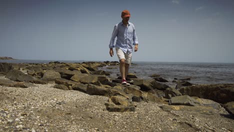 man sits on the rocks on the beach front then gets up and walks off past the camera.