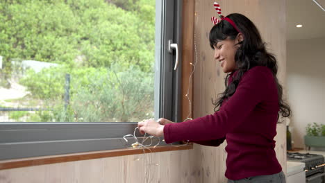 woman decorating window with christmas lights, wearing festive headband at home
