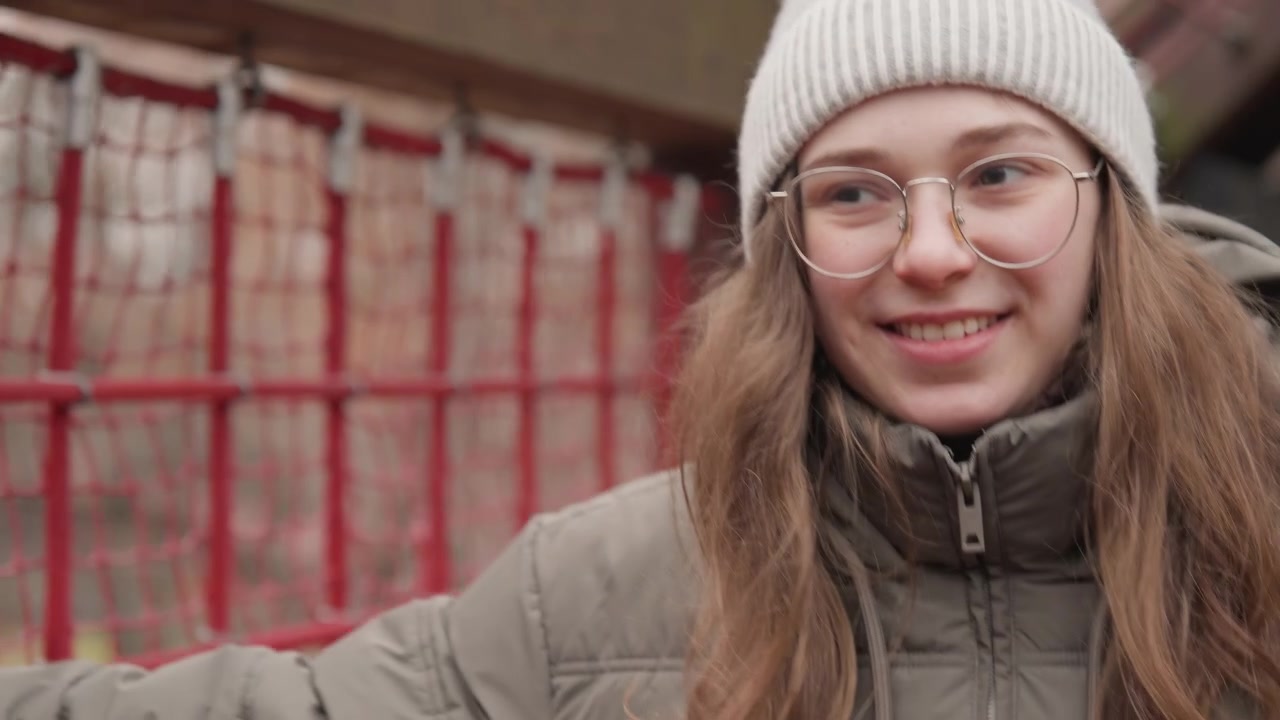Donna bianca con berretto di lana sorridente, scena rustica all'aperto vicino a una recinzione rossa e una struttura in legno, piumino e occhiali, atmosfera giovanile da studente, energia amichevole e locale, luce soffusa e nuvolosa, spontanea.