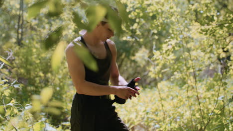 young man stands amid sunlit forest foliage wearing headband and fingerless glove, reaching forward as branch tip comes into sharp focus in blurred foreground while he adjusts glove strap to move on