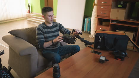 young man in striped sweater preparing camera gear, checking camera equipment on wooden table, organizing tools for shooting in cozy living room, readying filming equipment for production