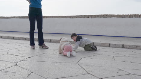 dos bebés se arrastran por la calle de piedra mientras la madre está de pie y mira con alegría