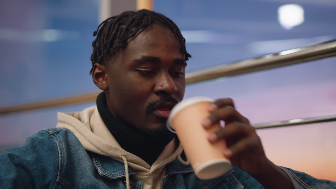 Close up of young man with braided hair wearing beige hoodie and denim jacket sitting quietly indoors during soft sunset, eyes gently lowered, surrounded by warm evening tones and reflective mood lighting