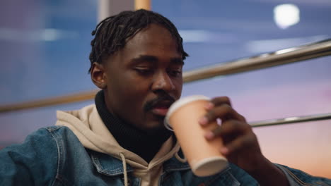 close up of young man with braided hair wearing beige hoodie and denim jacket sitting quietly indoors during soft sunset, eyes gently lowered, surrounded by warm evening tones and reflective mood lighting