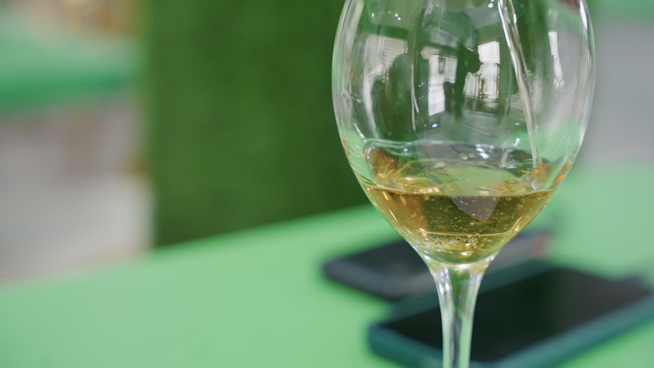 Bicchiere di vino e telefono su un tavolo verde, che indugiano dopo un appuntamento romantico in patio, piccola quantità di vino bianco in un bicchiere a stelo, luce solare soffusa che crea un bokeh caldo, atmosfera da caffè informale, umore intimo e rilassato.