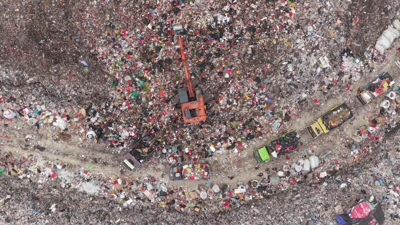 Veduta aerea dall'alto che mostra un convoglio di camion della spazzatura che si muove attraverso un'enorme discarica a cielo aperto, con un escavatore che sposta enormi pile di plastica e altri rifiuti non riciclabili.