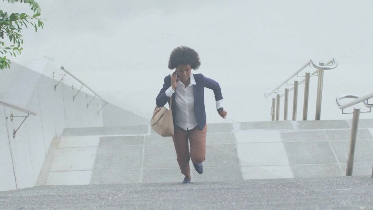 Businesswoman climbing outdoor stairs, speaking on phone after checking watch, quickening pace