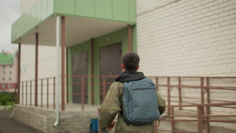 close up rear view of kid carrying backpack glancing sideways while running toward building entrance with blurred residential structures in background and green surroundings under cloudy sky