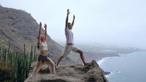 a man and woman stand on a cliff, raise their hands, and breathe in the sea air during yoga, their focus on the expansive ocean