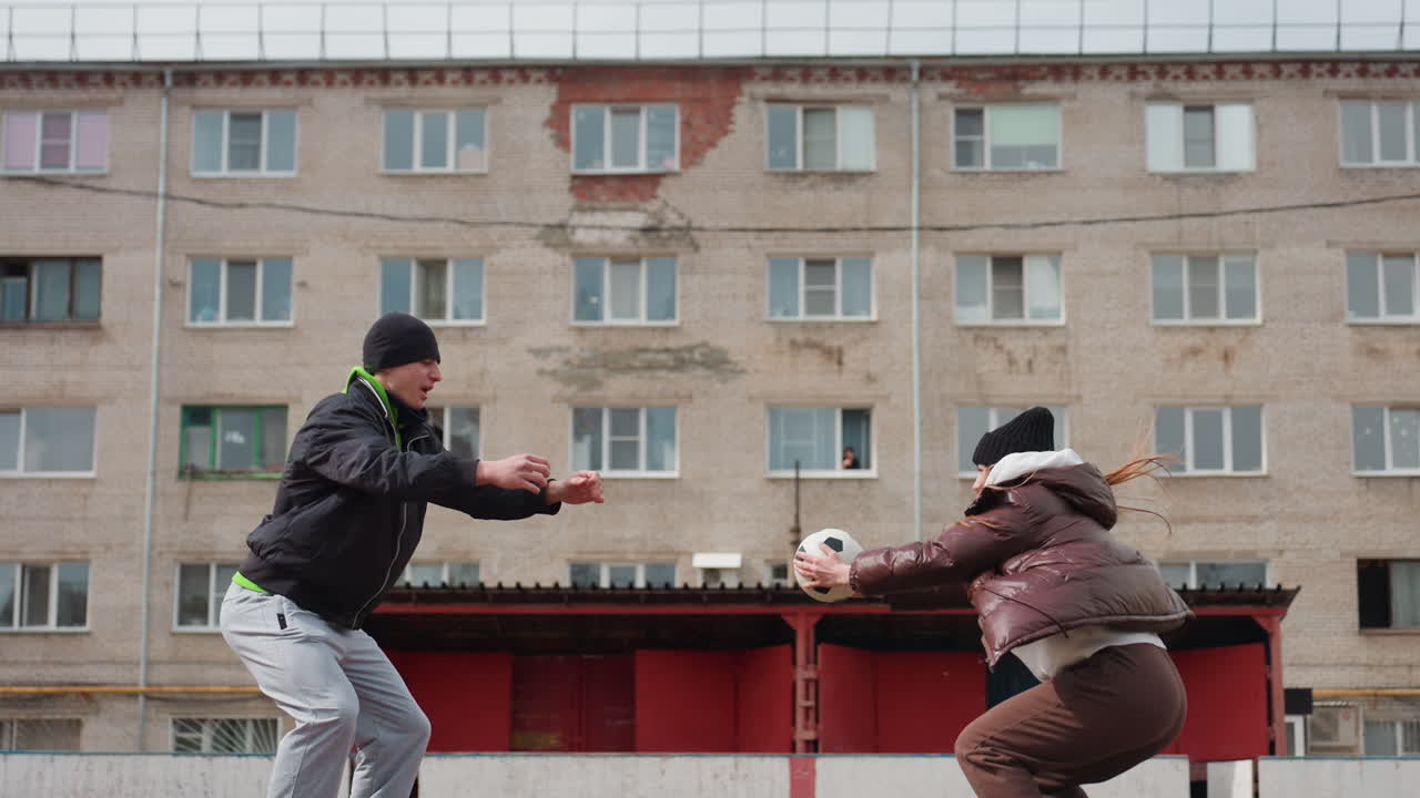 백인 남성 강사와 노년 여성 선수가 도시 안뜰에서 공을 주고받으며 가까이에서 건네고 던지는 모습, 아파트 단지가 배경에 있고, 방한 재킷과 운동화를 착용, 부드러운 코칭 순간
