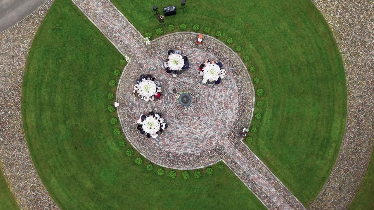 Video de dron que captura una elegante reunión al aire libre en un jardín paisajístico con invitados sentados en mesas redondas sobre un patio circular de ladrillo. Césped verde exuberante y senderos formales rodean el evento.