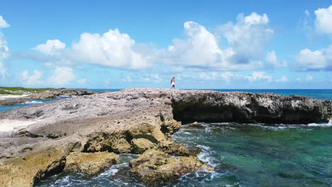 aerial of young girl in white dress cautiously walking on sharp rocks on coastline of cozumel mexico on sunny summer day