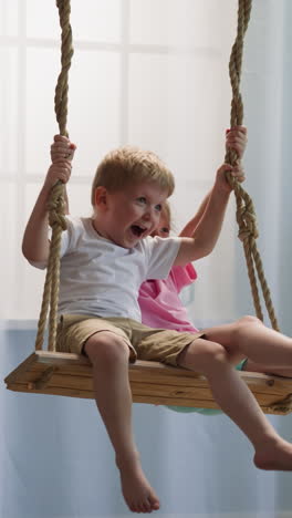 happy brother and sister sit on swing against big window at home. laughing children relax joyfully spending time together in cozy apartment