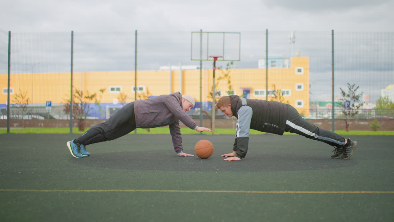 outdoor basketball court partners plank challenge two adults performing synchronized plank and pushup moves with basketball between them casual athletic clothing fenced urban court overcast sky
